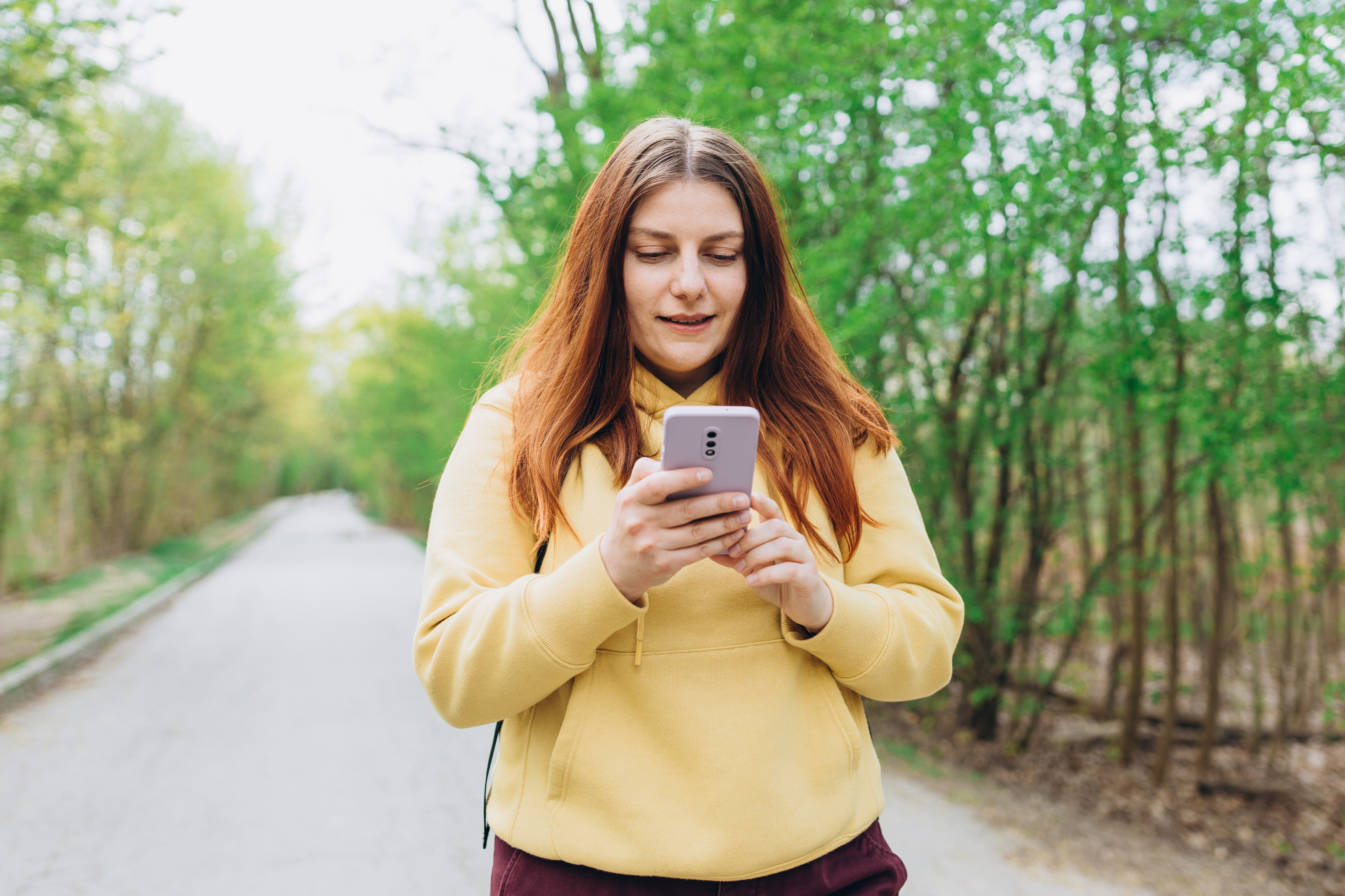 Frau mit langen, roten Haaren und gelbem Pullover auf einem Waldweg gehend, schaut auf ihr Handy